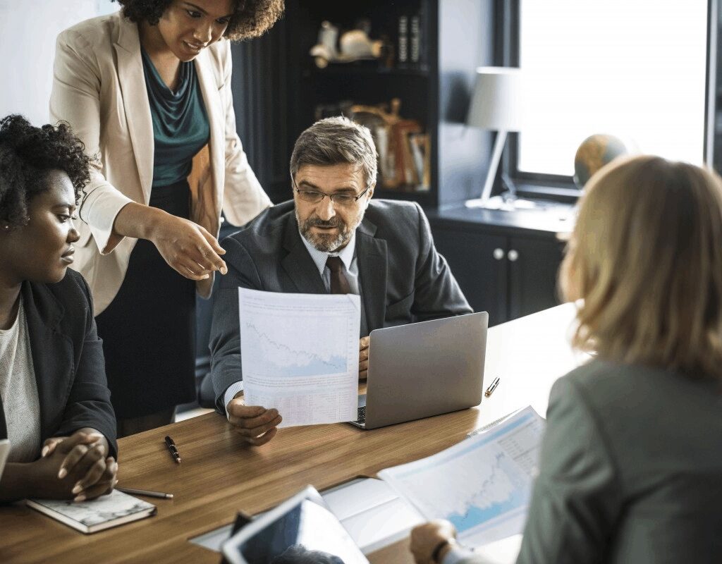 In a London meeting room, a group of professionals is absorbed in discussing financial documents. A woman highlights graphs while a man with glasses listens attentively. Two others study the papers, considering how SEO services in London could enhance their strategy. A laptop sits open on the table.